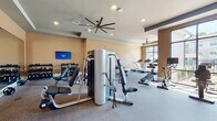 This fitness center in Memphis, TN offers a comprehensive workout environment designed to accommodate a wide range of exercise routines. The space is organized across a single floor and divided into distinct functional zones, each thoughtfully arranged to maximize usability.

The strength training area features a multi-station cable machine, a power rack, adjustable benches, medicine ball storage, and an extensive dumbbell rack with a full range of weights, all mounted along warm-toned beige walls. A wall-mounted flat-screen TV provides entertainment for those in this section.

The cardio zone is situated near a large floor-to-ceiling window, flooding the area with natural light and providing outdoor views of the surrounding property. Treadmills, stationary bikes, and an elliptical machine are positioned to take advantage of this bright, open atmosphere. Ceiling fans with wide blade spans are installed throughout, promoting air circulation and comfort during workouts.

A separate glass-walled room with bold graphic lettering on the panels provides a dedicated space for stretching, functional training, or small group exercise. Recessed lighting and carpet flooring throughout contribute to a clean, polished aesthetic that balances functionality with a welcoming ambiance.