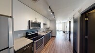 This contemporary 2-bedroom, 2-bathroom unit in Austin, TX (78705) offers a well-designed single-floor layout with modern finishes throughout. The galley-style kitchen is outfitted with stainless steel appliances — including an electric range, over-the-range microwave, dishwasher, and refrigerator — complemented by white upper cabinetry, dark lower cabinets, light quartz countertops, and a stainless steel sink. Track lighting illuminates the kitchen, which flows seamlessly into the living area via warm wood-look vinyl flooring. The living room is a standout space, featuring a large floor-to-ceiling window that floods the room with natural light and frames sweeping views of the Austin skyline and surrounding greenery. An exposed concrete ceiling adds an industrial character to the area. Each bedroom includes built-in closets with wire shelving, providing functional storage. The primary bedroom features dual closets for added convenience. Both bathrooms are finished with white tile flooring, dark vanity cabinetry, and clean white walls, maintaining the unit's consistent modern aesthetic. Dark-framed doors throughout contribute to a sleek, cohesive design. This unit combines urban functionality with contemporary style in a prime Austin location.