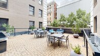 Located at 145 Granby Street in the heart of downtown Norfolk, Virginia, this outdoor terrace offers a well-appointed open-air retreat surrounded by the architecture of the city. The terrace is finished with a herringbone-patterned brick paver surface that adds a classic, urban elegance to the space. Enclosed on multiple sides by the warm tan brick façade of the building, the area feels both sheltered and connected to the surrounding cityscape.

The terrace is thoughtfully furnished to accommodate a variety of uses. Multiple dining-height tables with coordinating chairs are arranged across the paved surface, providing ample seating for outdoor meals or casual gatherings. A dedicated lounge area features plush blue cushioned sofas and a low coffee table, creating a comfortable spot for relaxation. Grilling stations are also available, making the space ideal for outdoor cooking and entertaining.

Greenery is woven throughout the terrace, with mature trees lining the perimeter wall and large rounded planters holding ornamental trees and shrubs. The plantings soften the urban setting and provide a sense of privacy. Views of neighboring downtown Norfolk buildings frame the space, reinforcing its vibrant city-center location. The terrace is accessed via a covered entryway and features railing-defined zones that add structure and definition to the open layout.