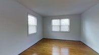 This single-floor space offers a clean, understated aesthetic defined by its warm hardwood flooring and crisp white walls. The main living area is bright and open, benefiting from multiple windows fitted with horizontal blinds that allow for adjustable natural light throughout the day. The windows frame views of the surrounding exterior, bringing a sense of connection to the outdoors.

The flooring throughout features classic hardwood in a rich honey tone, adding warmth and character to the otherwise minimal interior. White baseboards run along the perimeter of the room, providing a clean transition between the walls and floors and contributing to the space's tidy, well-maintained appearance.

The smooth white ceiling and neutral color palette create a versatile backdrop well-suited to a variety of furnishing styles. Baseboard heating units are visible beneath the windows, offering a practical and unobtrusive heating solution. The layout feels open and functional, with the simplicity of the design lending itself to easy customization. Overall, the space presents a bright, welcoming environment characterized by its natural light, classic hardwood floors, and clean, straightforward finishes.