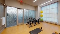 Located at 2250 Hawkins St. in Charlotte's South End neighborhood, this single-floor fitness studio is thoughtfully designed to support a variety of workout modalities. The space features a durable cork-style flooring that provides comfort and impact absorption for floor-based exercises. Natural light pours in through large windows dressed with sheer curtains, creating a bright and energizing atmosphere throughout the room.

The studio is equipped with a row of stationary cycling bikes, ideal for spin-style workouts, alongside multiple exercise mats and yoga blocks for stretching, yoga, and mat-based training. A dedicated barre rail runs along one mirrored wall, offering support for ballet-inspired fitness routines, while the full-length mirror panel allows users to monitor form and technique. A collection of stability balls and kettlebells rounds out the available equipment.

A wall-mounted flat-screen television serves as a focal point on one end of the room, complemented by a sleek linear display beneath it, offering an audiovisual element for guided workouts or classes. Overhead linear LED lighting ensures consistent, even illumination throughout. Decorative frosted glass panels with geometric patterns divide the space and add an elegant architectural touch. The studio's clean, neutral color palette and well-organized layout create a focused and motivating environment for fitness activity.