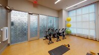 Located at 2250 Hawkins St. in Charlotte's South End neighborhood, this single-floor fitness studio is thoughtfully designed to support a variety of workout modalities. The space features a durable cork-style flooring that provides comfort and impact absorption for floor-based exercises. Natural light pours in through large windows dressed with sheer curtains, creating a bright and energizing atmosphere throughout the room.

The studio is equipped with a row of stationary cycling bikes, ideal for spin-style workouts, alongside multiple exercise mats and yoga blocks for stretching, yoga, and mat-based training. A dedicated barre rail runs along one mirrored wall, offering support for ballet-inspired fitness routines, while the full-length mirror panel allows users to monitor form and technique. A collection of stability balls and kettlebells rounds out the available equipment.

A wall-mounted flat-screen television serves as a focal point on one end of the room, complemented by a sleek linear display beneath it, offering an audiovisual element for guided workouts or classes. Overhead linear LED lighting ensures consistent, even illumination throughout. Decorative frosted glass panels with geometric patterns divide the space and add an elegant architectural touch. The studio's clean, neutral color palette and well-organized layout create a focused and motivating environment for fitness activity.