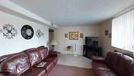 This single-floor home offers a well-organized layout across several functional living spaces. The living room serves as the central gathering area, finished with neutral carpet and a textured ceiling. Leather sofas are arranged around a dark media console housing a flat-screen TV and an electric fireplace insert, creating a warm focal point. Vertical blinds on a large window allow natural light to filter in, brightening the room. Framed floral artwork decorates the walls, adding a homey touch.

The kitchen is efficiently designed with warm oak cabinetry providing ample upper and lower storage. White appliances, including a refrigerator, dishwasher, and electric range, are neatly integrated. Tile flooring runs throughout, contrasting with the laminate countertops that offer generous prep space.

Adjacent to the kitchen, the dining area features a glass-top table beneath a ceiling fan with a light fixture. A decorative wrought-iron wall piece adds character, while red-checked curtains on a nearby window introduce a classic, welcoming feel. A wooden spindle divider partially separates this space from the neighboring area.

The bedroom is carpeted and comfortably sized, featuring a window with deep-toned curtains, matching nightstands, and a full mirrored sliding-door closet that enhances the sense of space and reflects natural light effectively.