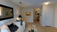 This single-floor residence presents a thoughtfully designed open-concept layout that promotes a natural flow between living spaces. The living room serves as the central hub, featuring neutral-toned walls and light carpeting that create a warm, inviting atmosphere. A comfortable white sofa anchors the space, complemented by accent lighting from a sleek black table lamp, adding a soft, ambient glow to the room.

A distinctive architectural pass-through window connects the living area to the kitchen, offering both visual connectivity and practicality. The kitchen, partially visible through the pass-through, features light cabinetry and appears bright and functional.

A wide opening leads from the living room into a dedicated dining area, where an elegant multi-light pendant chandelier hangs above a dark-framed dining set, creating a refined contrast against the neutral surroundings. Wall art and a mirror add decorative character to this space. The consistent use of light, neutral tones throughout — from the carpeting to the walls — contributes to a cohesive, airy aesthetic across the entire residence.