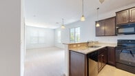 This single-floor residence offers a thoughtfully designed open-concept layout that seamlessly connects the kitchen and living area. The kitchen is a standout feature, showcasing rich dark-wood cabinetry, granite countertops, and a full suite of black appliances including a range, built-in microwave, dishwasher, and refrigerator. A peninsula with pendant lighting overhead serves as both a functional workspace and a natural divider between the kitchen and the expansive carpeted living area, which benefits from multiple windows that invite abundant natural light throughout the day. The bedrooms are finished with soft, neutral-toned carpet and feature window blinds that allow for flexible light control. The primary bathroom is equipped with a wide vanity featuring dark-wood cabinetry, a white countertop, a large mirror, and a tub-shower combination, while a second bathroom offers a walk-in shower stall and coordinating finishes. Tile flooring runs through the bathrooms and kitchen areas, providing a practical contrast to the plush carpet in the living and sleeping spaces. A dedicated laundry room with full-size top-load washer and dryer adds significant everyday convenience. Crisp white walls and consistent finishes throughout lend the space a clean, cohesive aesthetic.