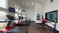 Located at 1514 Ennis Joslin Rd in Corpus Christi, Texas, this single-floor fitness center is designed to support a comprehensive workout experience. The space is defined by its soaring vaulted ceilings and an expansive bank of large windows along one wall, flooding the interior with abundant natural light while offering outdoor views of a pool area and surrounding palm trees — a fitting backdrop for exercise in a coastal Texas setting.

The cardio zone is well-appointed with multiple Freemotion treadmills, an elliptical trainer, and a recumbent bike, each equipped with individual monitors. Wall-mounted flat-screen televisions are positioned above the windows for additional entertainment during workouts. The strength training area features a full power rack with Olympic weight plates, a flat/adjustable bench, a dumbbell rack, and kettlebells, catering to a range of resistance training needs. Large floor-to-ceiling mirrors line one side of the strength zone, enhancing the sense of space and allowing users to monitor their form.

A dedicated functional training wall displays foam rollers, medicine balls, and resistance accessories for flexibility and conditioning work. The flooring throughout is a durable dark surface suited for high-traffic athletic use. The space also includes safety amenities such as a mounted fire extinguisher and clearly marked emergency exits, ensuring a safe and functional environment for all users.