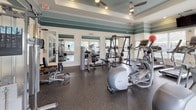 This single-story amenity space combines a professional business center with a fully equipped fitness facility. The entrance corridor sets a polished tone with light tile flooring laid on the diagonal, crown molding, framed artwork, and a wall-mounted water fountain, leading through a glass-paneled door into the interior.

The business center features two distinct workspaces. The main room offers multiple computer workstations with dark wood desks featuring granite-style surfaces, ergonomic chairs, and wall-mounted artwork against warm taupe accent walls. A second, more private workstation area provides a focused workspace with carpeted flooring and natural light filtering through a window dressed with elegant drape panels.

The fitness center is a standout space, distinguished by a tray ceiling accented with teal and cream striping, recessed lighting, and ceiling fans. Large windows along the perimeter flood the room with natural light while offering outdoor views. The rubber-floored gym is well-equipped with treadmills, ellipticals, strength training machines, free weights, and benches. Full-length mirrors line one wall, enhancing the sense of openness. Together, these amenities form a cohesive, thoughtfully designed facility.