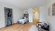 This single-floor studio is designed with a modern, minimalist aesthetic that maximizes its open layout. The main living area seamlessly integrates a sleeping zone and a sitting area, creating a functional and cohesive space. Warm-toned hardwood flooring runs throughout, adding a sense of warmth that contrasts pleasantly with the crisp white walls and ceiling.

The sleeping area is anchored by a bed fitted with a charcoal upholstered headboard, positioned against a partial wall divider that subtly defines the space without fully enclosing it. Decorative animal skull wall art adds a subtle stylistic accent above the headboard. A transparent side table provides practical surface space without visually cluttering the room.

The sitting area features a cushioned armchair paired with a dark side table, set atop a light-toned area rug that grounds the zone. Recessed lighting in the entry corridor provides additional illumination, while the entryway leads to a secure entry door equipped with a keypad lock. The overall ambiance is clean, calm, and well-organized, offering a straightforward and efficiently designed living space.