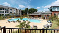 This thoughtfully designed outdoor amenity space serves as the centerpiece of a modern residential community, combining resort-style recreation with refined landscaping and comfortable gathering areas. The focal point is a generously sized freeform swimming pool with crystal-clear water, surrounded by a spacious tiled sundeck furnished with chaise lounges and seating clusters ideal for relaxing or socializing. A dedicated wading or entry area provides a gradual water entry, adding both functionality and visual appeal to the pool design. Bordering the pool, black iron fencing defines the space while maintaining open sightlines to the surrounding grounds. A covered pavilion anchored by brick columns provides a shaded outdoor lounge area outfitted with plush seating and a low coffee table, offering a comfortable retreat that overlooks the natural landscape. The surrounding grounds are professionally landscaped with ornamental grasses, native plantings, young trees, and pine straw beds that lend a natural, wooded aesthetic. The community buildings — featuring a mix of brick, siding, and teal-accented architectural details — frame the outdoor space and include a distinctive cupola-topped clubhouse. The overall ambiance is vibrant yet relaxed, well-suited for year-round outdoor enjoyment.