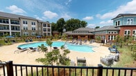 This thoughtfully designed outdoor amenity space serves as the centerpiece of a modern residential community, combining resort-style recreation with refined landscaping and comfortable gathering areas. The focal point is a generously sized freeform swimming pool with crystal-clear water, surrounded by a spacious tiled sundeck furnished with chaise lounges and seating clusters ideal for relaxing or socializing. A dedicated wading or entry area provides a gradual water entry, adding both functionality and visual appeal to the pool design. Bordering the pool, black iron fencing defines the space while maintaining open sightlines to the surrounding grounds. A covered pavilion anchored by brick columns provides a shaded outdoor lounge area outfitted with plush seating and a low coffee table, offering a comfortable retreat that overlooks the natural landscape. The surrounding grounds are professionally landscaped with ornamental grasses, native plantings, young trees, and pine straw beds that lend a natural, wooded aesthetic. The community buildings — featuring a mix of brick, siding, and teal-accented architectural details — frame the outdoor space and include a distinctive cupola-topped clubhouse. The overall ambiance is vibrant yet relaxed, well-suited for year-round outdoor enjoyment.