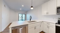 Located at 8707 Shawnee Mission Pkwy in Merriam, Kansas, this one-bedroom, one-bathroom unit offers a thoughtfully designed single-floor layout with a clean, modern aesthetic throughout. The open-concept kitchen and living area serves as the heart of the space, anchored by crisp white shaker cabinetry, quartz countertops, and a full suite of stainless steel Whirlpool appliances including a refrigerator, electric range, over-the-range microwave, and dishwasher. A peninsula with a matte black faucet and undermount sink adds both function and style, while a pendant light overhead provides warm accent lighting. Wood-look flooring flows throughout the living area, transitioning to a richer, darker tone in the bedroom. Sliding glass doors lead from the living space to an outdoor area, bringing in natural light. The bedroom is generously proportioned, featuring a ceiling fan, natural light from a window, and a dedicated closet. The bathroom is finished with white shaker vanity cabinetry, a tiled tub/shower surround, and coordinating matte black fixtures. An in-unit laundry room houses a full-size washer and dryer alongside a water heater and HVAC system, completing this efficiently designed and well-appointed home.