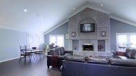 This single-floor home showcases an open-concept living and dining area anchored by a dramatic floor-to-ceiling brick fireplace wall, which serves as the room's defining architectural centerpiece. The brick surround features a distinctive arched detail above the fireplace opening, with a mounted flat-screen television integrated seamlessly into the design. Framed artwork flanks the fireplace, adding a personal touch to the bold masonry backdrop.

The vaulted ceiling rises steeply, following the roofline to create an impressive sense of vertical space and airiness throughout the room. Recessed lighting is embedded along the ceiling slope, providing even, ambient illumination. The walls are painted in a cool slate blue, complementing the warm tones of the brick and the rich dark hardwood flooring that runs throughout.

The living area is furnished with deep, dark leather sofas arranged to face the fireplace, creating an inviting focal point. Adjacent to the living space, a casual dining area accommodates a round table and wrought-iron chairs. Large windows dressed with plantation shutters line the far wall, allowing natural light to filter through while maintaining privacy.