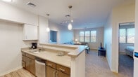 This single-floor unit is designed with a functional, modern layout that balances comfort and practicality. The kitchen serves as a central hub, featuring light wood-grain flooring, medium-toned wood cabinetry, light countertops, stainless steel appliances including a refrigerator and dishwasher, and an undermount sink. A peninsula with pendant lighting connects the kitchen to the open living area, which is finished with plush carpeting and a wide window that brings in generous natural light and outdoor views.

The living area offers a comfortable setting with a sofa, armchair, and coffee table arranged around a television. A separate seating nook with two chairs and a side table provides an additional relaxed retreat, also benefiting from natural light through a window with blinds.

Both bedrooms are carpeted and feature windows that allow natural light in. Each is spacious enough to accommodate a queen-sized bed and additional furnishings. One bedroom includes a dresser with a wall mirror and direct access to a hallway leading to the bathroom.

The two bathrooms are finished with neutral tile flooring and feature dark wood vanities with light countertops. Both include accessibility-focused grab bars, with one offering a shower-tub combination and the other a walk-in shower. The overall ambiance throughout is clean, neutral, and quietly contemporary.