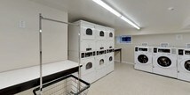 This shared laundry facility is a practical, well-maintained space designed to accommodate multiple users simultaneously. The layout is open and organized, with light-toned walls and durable speckled flooring throughout that contribute to a clean and hygienic environment. Bright fluorescent lighting runs along the ceilings, ensuring the space is well-lit at all times.

The laundry area is equipped with a generous number of commercial-grade front-loading washing machines arranged in rows along one side, complemented by multiple stacked dryer units on the opposite side. Each machine features digital controls for ease of use. Folding tables with a dark-edged finish provide ample surface area for sorting and folding laundry, and wire laundry carts are available for convenient transport.

An instructional and payment information board is mounted on the wall, offering guidance for users. A trash bin and cleaning supplies are also accessible within the space. Small windows near the ceiling allow a modest amount of natural light to supplement the overhead lighting.

Adjacent to the main laundry area is a waiting room featuring a row of black chairs along the wall, a centrally located structural column, and a doormat at the entrance. A fire extinguisher and exit signage are prominently placed, reflecting attention to safety. A door leads further into the facility, completing this functional and user-friendly communal space.