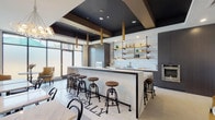 This contemporary single-floor space is thoughtfully designed to blend a fully equipped kitchen with a sophisticated lounge and dining area. At the heart of the space sits a striking large-format marble-topped island, offering bar-height seating with industrial-style adjustable stools featuring warm wooden seats. The kitchen wall showcases a clean white subway tile backsplash, complemented by dark wood cabinetry and open pipe-mounted wood shelving displaying glassware and decorative accents. Stainless steel appliances — including a wall oven, microwave, warming drawer, beverage cooler, and sink — provide a full suite of culinary functionality. The flooring transitions between large-format light gray tile and a patterned hexagonal mosaic tile beneath the island area, adding visual interest and defining zones within the open layout. The dining area features tufted gray banquette seating alongside marble-topped tables and classic bistro chairs, while a cozy seating nook with mustard yellow accent chairs provides an intimate retreat. Steel-framed glass partitions delineate spaces while maintaining openness and light. Overhead, a dramatic dark-painted ceiling with gold cylindrical pendants and a globe chandelier adds depth and an upscale ambiance. Large frosted windows allow ample natural light throughout.