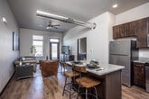 This single-floor unit showcases a thoughtfully designed open-concept layout with distinct industrial-modern character. An exposed spiral ductwork system runs prominently across the ceiling of the main living area, complemented by a ceiling fan and recessed lighting that together create a loft-like atmosphere. Rich hardwood-style flooring flows consistently throughout the space.

The kitchen is well-appointed with dark espresso cabinetry, stainless steel appliances including a refrigerator, range, microwave, and dishwasher, plus a central island with light quartz-style countertops offering bar seating. Open shelving adds both function and visual interest to the kitchen walls.

The living area provides generous space for seating arrangements and benefits from natural light through large windows that face an outdoor balcony. The bedroom is comfortably sized with tall, narrow windows offering outdoor views and ample natural light, accompanied by hardwood-style flooring and neutral tones throughout.

The bathroom features a dark-framed glass door, a light quartz vanity countertop, and clean modern finishes. A dedicated laundry closet houses a stacked full-size washer and dryer, adding everyday convenience to the unit.