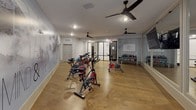 This well-appointed fitness studio is thoughtfully designed to support a comprehensive workout experience across its single-floor layout. The centerpiece of the space is a spacious spin cycle room outfitted with multiple stationary bikes arranged for group cycling sessions. A dramatic floor-to-ceiling mountain mural dominates one wall, accompanied by the motivational phrase "Quiet the Mind & the Soul Will Speak," creating an immersive and inspiring atmosphere. Large wall-to-wall mirrors reflect the room, amplifying the sense of space, while a wall-mounted flat-screen television adds a modern amenity. Ceiling fans ensure comfortable airflow during intense sessions, and warm wood-toned flooring runs throughout, lending a clean, contemporary feel. Natural light filters in through shuttered windows, brightening the interior. An adjacent area connects to a dedicated strength and functional training zone, equipped with kettlebells, weight racks, and additional cardio machines. Stability balls and smaller fitness accessories are neatly stored along the walls. Black-framed glass partitions elegantly divide the zones while maintaining visual openness throughout the studio. The overall ambiance is modern, motivating, and professionally designed to encourage active, focused fitness routines.