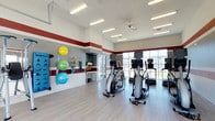 This fitness center offers a clean, well-organized space designed to support a full range of workout routines. The room features high ceilings accented by a bold red horizontal stripe along the upper walls, giving the space a dynamic and energetic feel. Large floor-to-ceiling windows along one wall allow abundant natural light to fill the room while offering views to the exterior, creating an open and inviting atmosphere. The flooring is a light-toned hardwood-style surface that is both durable and easy to maintain. A ceiling fan overhead aids in air circulation during workouts. The cardio area is equipped with multiple elliptical trainers and treadmills, arranged to maximize floor space and usability. Along the opposite wall, a dedicated functional fitness zone features a colorful array of exercise balls — including stability balls and medicine balls — mounted on a wall rack, alongside resistance bands and other accessories. Bright recessed and overhead fluorescent lighting ensures the entire space is well-lit at all times. A small built-in counter or storage area is accessible through an interior doorway, adding a practical element to the facility. Overall, the fitness center presents a bright, spacious, and thoughtfully arranged environment suitable for a variety of fitness activities.