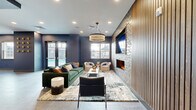 Located at 120 Matheson Ave in Charlotte, NC, this thoughtfully designed single-floor space seamlessly combines a communal kitchen and living room into one open-concept environment. The living area makes a strong visual statement with a full-height stacked stone fireplace wall flanked by warm wood slat paneling, a wall-mounted TV, and a tiered chandelier that anchors the seating area below. A plush emerald green sofa, curved cream accent chairs, and a layered area rug create an inviting and curated atmosphere. Natural light filters in through multiple windows, offering views of the surrounding property. The kitchen area is equally striking, centered around a large island with a white quartz countertop and sculptural arch-back barstools. Dark cabinetry lines the perimeter, complemented by a vibrant multicolored mosaic tile backsplash and warm pendant lighting. A bold gallery wall featuring eclectic framed artwork adds a creative, urban energy to the space. Large-format tile flooring runs throughout, tying both zones together cohesively. The overall design balances contemporary sophistication with an energetic, artistic character, making it a compelling and functional communal gathering space.