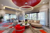 This thoughtfully designed community lounge and co-working space seamlessly blends social and professional functionality within a single, open-plan floor. The most striking architectural feature is the dramatic circular red ceiling medallion, beneath which a large halo-shaped LED ring chandelier provides a warm, ambient glow that anchors the lounge area below. The ceiling transitions to a contrasting textured concrete-finish tray in the conference zone, creating distinct yet cohesive environments within the space.

The lounge area is furnished with a curved sectional sofa, bold red accent chairs, and a round coffee table, all set atop a patterned modular carpet in grey, red, and blue tones that adds visual energy to the floor. A built-in wet bar with decorative tile backsplash and appliances lines one wall, making the space well-suited for gatherings and events.

Adjacent to the lounge, a dedicated conference area features a long rectangular table surrounded by ergonomic task chairs, illuminated by a linear pendant light. A wall-mounted display screen near the entry supports presentations and digital signage.

Floor-to-ceiling windows and glass doors line the perimeter, flooding the interior with natural light and offering views of the surrounding outdoor environment. Sheer white drapery softens the window walls throughout, enhancing the polished, contemporary ambiance of the overall space.