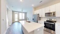 This well-designed 1-bedroom, 1-bathroom residence offers a thoughtful single-floor layout that maximizes functionality and comfort. The open-concept kitchen and living area serves as the heart of the home, featuring a large center island with a white quartz countertop, an undermount sink with a brushed gold faucet, and ample seating potential. The kitchen is outfitted with crisp white shaker-style cabinetry, a subway tile backsplash, and a full suite of stainless steel appliances, including a French door refrigerator with ice maker, electric range, and built-in microwave. Wood-look flooring extends throughout the main living area, complementing the neutral color palette and recessed lighting overhead. A ceiling fan anchors the living space, which opens to a private balcony through a glass-paneled door, allowing natural light to pour in. The bedroom is carpeted for warmth and comfort, with large windows framing outdoor views and a walk-in closet equipped with wire shelving for organized storage. The bathroom features light tile flooring, a white vanity with quartz countertops, and clean, modern fixtures. A dedicated laundry area rounds out the practical amenities of this efficiently designed residence.