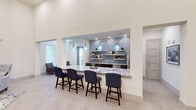 This single-floor space is designed with a contemporary aesthetic, combining functionality and style across several distinct areas. Soaring ceilings create an expansive, airy atmosphere throughout, enhanced by large windows and a skylight that flood the interior with natural light.

The centerpiece is a generous bar-style counter finished in striking granite, lined with upholstered navy bar stools and illuminated by a trio of artisan blue glass pendant lights. Behind the counter, a well-appointed kitchenette features dark cabinetry, stainless steel appliances, a double sink, and open floating shelves set against a decorative blue mosaic tile backsplash that adds texture and visual interest.

A sitting nook near the entrance offers a tranquil retreat, with two armchairs, a bistro table, and views to the exterior through slatted blinds, all beneath a recessed skylight. A lounge area anchored by a bold navy accent wall showcases a mounted flat-screen television, decorative gold sunburst mirrors, and a tufted wingback chair atop a patterned area rug. Large-format porcelain tile flooring flows seamlessly throughout, unifying the space. The open overhead view reveals a thoughtfully arranged layout with multiple seating configurations suitable for gatherings or events.