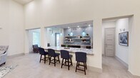 This single-floor space is designed with a contemporary aesthetic, combining functionality and style across several distinct areas. Soaring ceilings create an expansive, airy atmosphere throughout, enhanced by large windows and a skylight that flood the interior with natural light.

The centerpiece is a generous bar-style counter finished in striking granite, lined with upholstered navy bar stools and illuminated by a trio of artisan blue glass pendant lights. Behind the counter, a well-appointed kitchenette features dark cabinetry, stainless steel appliances, a double sink, and open floating shelves set against a decorative blue mosaic tile backsplash that adds texture and visual interest.

A sitting nook near the entrance offers a tranquil retreat, with two armchairs, a bistro table, and views to the exterior through slatted blinds, all beneath a recessed skylight. A lounge area anchored by a bold navy accent wall showcases a mounted flat-screen television, decorative gold sunburst mirrors, and a tufted wingback chair atop a patterned area rug. Large-format porcelain tile flooring flows seamlessly throughout, unifying the space. The open overhead view reveals a thoughtfully arranged layout with multiple seating configurations suitable for gatherings or events.