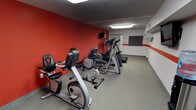 This fitness center is designed to offer a functional and energizing workout environment. The space is defined by its striking red accent walls, which contrast with the neutral gray tones of the flooring and ceiling, creating a dynamic and motivating atmosphere. The room is outfitted with a selection of cardio equipment, including stationary recumbent bikes, upright bikes, and elliptical trainers, arranged in an organized row to maximize the use of floor space. Overhead fluorescent lighting provides bright, even illumination throughout the room, ensuring visibility across all workout areas. A wall-mounted television is positioned to allow viewing while using the cardio machines, adding convenience for users during longer workout sessions. Toward the rear of the space, an open area with a dark padded mat provides room for stretching, bodyweight exercises, or additional training activities. The rubberized flooring is well-suited to a fitness environment, offering durability and comfort underfoot. Built-in shelving along one wall adds a practical storage element to the layout. Overall, the space is clean, purposeful, and thoughtfully arranged to support a range of fitness routines.