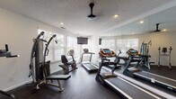 Located at 3170 Mapleleaf Dr in Lexington, Kentucky, this dedicated fitness center offers a comprehensive workout environment designed for both cardio and strength training. The space is thoughtfully laid out on a single floor, maximizing functionality and ease of movement between equipment. Multiple treadmills with integrated digital displays are arranged along one side of the room, complemented by a recumbent stationary bike, an elliptical trainer, and a multi-station cable strength machine, providing users with a full range of exercise options. A large wall-to-ceiling mirror spans one side of the room, visually expanding the space and allowing users to monitor their form during workouts. Natural light pours in through several white-shuttered windows that line the walls, offering pleasant views of surrounding greenery and creating an open, airy atmosphere. Recessed ceiling lighting and a ceiling fan ensure consistent illumination and comfort throughout the space. Wall-mounted flat-screen televisions provide entertainment options for users during cardio sessions. The flooring consists of dark rubberized material, ideal for high-traffic fitness use. Entry is through a set of elegant white French doors, adding a refined architectural touch. A sanitizing station is conveniently positioned near the entrance, promoting cleanliness and hygiene throughout the facility.