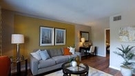 This two-floor residence offers a well-appointed living space with a design aesthetic that balances warmth and functionality. The living room serves as a central gathering area, featuring rich hardwood flooring that anchors the space and a textured ceiling that adds subtle architectural interest. A bold mustard-yellow accent wall creates a striking focal point, complemented by neutral gray tones on the surrounding walls for a balanced, contemporary palette.

The living room is furnished with a generously sized gray sofa adorned with decorative pillows in coordinating hues, including a vibrant orange accent pillow that echoes the room's warm color scheme. A round glass-topped coffee table with a dark wood base sits at the center, adding an elegant yet casual touch. Along one wall, a sleek console table holds a lamp and decorative accents, with a framed mirror above it enhancing the sense of depth. Two framed cityscape prints are displayed prominently on the accent wall, contributing to the room's curated, urban-inspired aesthetic. The overall ambiance is cozy and inviting, with warm lighting that highlights the space's carefully chosen design details.