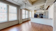 This two-bedroom, two-bathroom urban loft in Memphis, Tennessee offers a compelling blend of industrial character and modern finishes throughout its well-appointed single-floor layout. The open-concept living area immediately draws attention with its exposed overhead piping and ductwork, polished hardwood flooring, and generous windows fitted with blinds that invite abundant natural light while framing views of the surrounding cityscape. The kitchen is a functional centerpiece, featuring a large peninsula with granite countertops and an undermount sink, gray cabinetry, a mosaic tile backsplash, black appliances, and stylish pendant lighting suspended from the industrial ceiling. Both bedrooms are well-sized with clean white walls, hardwood floors, and multiple closets providing ample storage — one bedroom includes two separate closets and a ceiling fan. The two bathrooms are thoughtfully finished; one features a tub-shower combination with striking marble-patterned tile surround and a granite-topped vanity, while the other offers a walk-in glass-door shower with matching tile work. The consistent use of warm hardwood flooring, granite surfaces, and industrial-inspired architectural details throughout the unit creates a cohesive and stylish urban living environment in the heart of Memphis.