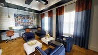 This stylish single-floor space makes a strong first impression with its bold, contemporary design aesthetic. The lounge area is anchored by a coordinated arrangement of deep navy blue armchairs and sofas surrounding sleek white marble-topped coffee tables, creating an inviting yet refined atmosphere. Gold and brass decorative accents — including vases, a textured bowl centerpiece, and pendant lighting — add warmth and visual contrast against the cool, dark color palette.

Floor-to-ceiling two-tone curtains in navy and burnt orange frame large windows that flood the interior with generous natural light, contributing to a bright and energizing ambiance. The ceiling features a distinctive coffered design with black geometric box details, lending architectural depth and a sense of elevated finish to the room.

Along one wall, a built-in workstation with dark countertops, leather-accented chairs, and computer monitors provides a dedicated functional area suited for business or productivity needs. A striking large-scale abstract artwork with metallic tones serves as a focal point near the workstation, reinforcing the space's curated, design-forward character.

Rich hardwood-style flooring grounds the space and complements the layered color scheme throughout. The overall ambiance is modern and polished, balancing comfort with sophistication.