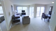 This single-floor space features an open-concept living area designed to maximize natural light and a sense of openness. The living room is generously sized, finished with neutral wall-to-wall carpeting that creates a clean, cohesive look throughout. Large windows along one wall allow abundant natural light to fill the space, offering views of surrounding greenery. A sliding glass door opens directly onto a private balcony, seamlessly connecting the interior to the outdoors and further enhancing the airy feel of the room.

The living area is arranged with comfortable seating, including a sofa and lounge chair, centered around a low-profile coffee table, with a flat-screen television mounted on a media console positioned near the windows. The layout allows for easy circulation and flexible furniture arrangement. The neutral color palette of soft grays and warm whites contributes to a calm, inviting atmosphere. Overall, the space presents a practical and comfortable layout with an emphasis on natural light, outdoor access, and an open, uncluttered aesthetic well-suited for everyday living.