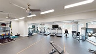This fitness center offers a generously sized, single-floor workout space designed to accommodate a wide range of exercise routines. The open floor plan is finished with durable rubber flooring marked with yellow lane lines, providing both functionality and a polished athletic aesthetic. Multiple ceiling fans and recessed panel lighting keep the environment comfortable and well-illuminated throughout.

The cardio zone is lined with treadmills, elliptical trainers, and a stationary bike, all positioned facing large windows that invite abundant natural light and offer views of the outdoors. A wall-mounted flat-screen television provides entertainment during workouts. A motivational typographic mural wraps along the main wall, adding an energizing visual element to the space.

The strength training area includes a multi-function cable machine, a dedicated free weights section with a full dumbbell rack, and an adjustable weight bench. A mirrored alcove along one wall creates a designated functional training zone equipped with suspension trainers and medicine balls, while also visually expanding the space.

Crown molding and clean white walls lend a refined architectural character uncommon in fitness facilities. Large glass doors along one side open to an outdoor area, further enhancing the sense of openness. The overall ambiance is bright, clean, and motivating — a professional-grade environment suited for a comprehensive fitness experience.