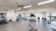 This fitness center offers a generously sized, single-floor workout space designed to accommodate a wide range of exercise routines. The open floor plan is finished with durable rubber flooring marked with yellow lane lines, providing both functionality and a polished athletic aesthetic. Multiple ceiling fans and recessed panel lighting keep the environment comfortable and well-illuminated throughout.

The cardio zone is lined with treadmills, elliptical trainers, and a stationary bike, all positioned facing large windows that invite abundant natural light and offer views of the outdoors. A wall-mounted flat-screen television provides entertainment during workouts. A motivational typographic mural wraps along the main wall, adding an energizing visual element to the space.

The strength training area includes a multi-function cable machine, a dedicated free weights section with a full dumbbell rack, and an adjustable weight bench. A mirrored alcove along one wall creates a designated functional training zone equipped with suspension trainers and medicine balls, while also visually expanding the space.

Crown molding and clean white walls lend a refined architectural character uncommon in fitness facilities. Large glass doors along one side open to an outdoor area, further enhancing the sense of openness. The overall ambiance is bright, clean, and motivating — a professional-grade environment suited for a comprehensive fitness experience.