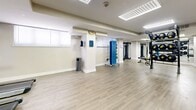 Located at 1131 W University Blvd W in Silver Spring, Maryland, this fitness center offers a thoughtfully designed, single-floor workout space that balances functionality with a clean, modern aesthetic. The open-plan layout is finished with light wood-style flooring and crisp white walls, creating a bright and welcoming environment conducive to exercise.

The space is well-lit by a combination of large recessed panel lights and skylights, ensuring even illumination throughout. Mirrored wall panels line one side of the room, visually expanding the space and providing useful visual feedback during workouts.

The fitness area is comprehensively equipped to support a wide range of training styles. Cardio equipment includes multiple treadmills positioned near a wall-mounted flat-screen television. Strength and functional training is supported by a multi-station cable machine, a medicine ball rack stocked with varied weights, a TRX suspension training system, a heavy battle rope, and an adjustable weight bench. A mobile foam roller and stretching board adds flexibility and recovery options.

Structural columns are integrated thoughtfully into the layout without impeding flow. Entry is through teal double doors, and the space includes a waste receptacle and informational signage. The overall ambiance is clean, professional, and purpose-built for an effective fitness experience.