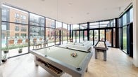 This thoughtfully designed single-floor amenity space offers residents a sophisticated and versatile environment for recreation and relaxation. The centerpiece of the space is a sun-drenched game room enclosed by dramatic floor-to-ceiling glass walls, allowing sweeping views of the outdoor pool courtyard and surrounding landscape. A full-size billiards table and shuffleboard table anchor the room, complemented by a plush white seating arrangement and a sculptural woven pendant light overhead. Adjacent to the game area is a comfortable lounge retreat, defined by soft shiplap-paneled walls in a calming sage tone and recessed ceilings with built-in lighting. This intimate seating area features a wall-mounted television, a linear fireplace framed in white tile, and an eclectic mix of upholstered sofas and artisanal wooden accent tables. The lounge is flanked on both sides by floor-to-ceiling windows that continue the connection to the outdoors. The kitchen is clean and contemporary, showcasing gray cabinetry, a striking blue-gray subway tile backsplash, a marble-topped island, herringbone hardwood flooring, and stainless steel appliances including a French door refrigerator. Pendant lighting adds warmth to the workspace. Together, these areas create a cohesive, design-forward environment ideal for both social gatherings and quiet leisure.