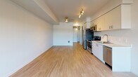 This contemporary single-floor unit showcases a clean, modern design throughout, highlighted by light wood-tone flooring and crisp white walls that create a bright, airy atmosphere. The open-concept layout seamlessly connects the kitchen and living area, maximizing the sense of space and functionality.

The kitchen is well-appointed with sleek two-tone cabinetry — white upper cabinets and grey lower cabinets — complemented by a stainless steel appliance suite that includes a refrigerator, electric range, over-the-range microwave, and dishwasher. Light countertops and a single-basin sink complete the efficient, modern cooking space.

The open living area benefits from a large window that invites natural light and offers outdoor views, along with recessed and track lighting overhead. Structural beam detailing on the ceiling adds a subtle architectural element to the space.

The bathroom features a shower-tub combination, a wall-mounted towel bar, overhead cabinetry for storage, and neutral tile flooring, all finished in a clean, contemporary style.

A dedicated utility area houses the electrical panel and a laundry hookup, adding practical convenience. Throughout the unit, the consistent use of light tones and modern finishes creates a cohesive, move-in-ready living environment.