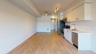 This contemporary single-floor unit showcases a clean, modern design throughout, highlighted by light wood-tone flooring and crisp white walls that create a bright, airy atmosphere. The open-concept layout seamlessly connects the kitchen and living area, maximizing the sense of space and functionality.

The kitchen is well-appointed with sleek two-tone cabinetry — white upper cabinets and grey lower cabinets — complemented by a stainless steel appliance suite that includes a refrigerator, electric range, over-the-range microwave, and dishwasher. Light countertops and a single-basin sink complete the efficient, modern cooking space.

The open living area benefits from a large window that invites natural light and offers outdoor views, along with recessed and track lighting overhead. Structural beam detailing on the ceiling adds a subtle architectural element to the space.

The bathroom features a shower-tub combination, a wall-mounted towel bar, overhead cabinetry for storage, and neutral tile flooring, all finished in a clean, contemporary style.

A dedicated utility area houses the electrical panel and a laundry hookup, adding practical convenience. Throughout the unit, the consistent use of light tones and modern finishes creates a cohesive, move-in-ready living environment.