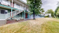 Located at 3498 Lovat Ave in Saanich, British Columbia, this multi-unit residential building offers a well-established presence within a tranquil neighbourhood setting. The two-storey structure is finished in a distinctive teal and grey exterior palette, with bold red entry doors adding a striking visual contrast along both levels. Each unit benefits from covered, railed walkways that run the full length of the building, providing sheltered access and a sense of continuity throughout the property. Upper-level units feature private balconies, while ground-floor units open onto their own covered porch areas — several of which have been personalized with seating and planters, reflecting a welcoming residential atmosphere. The building is flanked by generous, open lawn areas that extend across the property, offering ample green space framed by a diverse collection of mature trees, including large deciduous and coniferous specimens that provide shade and natural privacy. The landscaping contributes to a peaceful, park-like setting that enhances the overall ambiance of the property. Staircases at key points along the building connect the two levels efficiently. The surrounding grounds provide a comfortable outdoor environment, making this property a well-rounded residential option in the heart of Saanich.