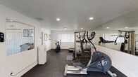 This single-level fitness center offers a comprehensive workout environment designed to accommodate a variety of exercise routines. The space is finished with clean white walls, recessed lighting, and durable dark rubber flooring throughout, creating a functional and motivating atmosphere.

The main workout area is outfitted with a range of cardio equipment, including a treadmill, an elliptical trainer, and an air resistance bike, providing options for diverse cardiovascular training. A large wall-mounted mirror runs along one side of the room, enhancing the sense of space while allowing users to monitor their form. A flat-screen TV is mounted on the adjacent wall for entertainment during workouts.

Strength and functional training are well supported, with a multi-function cable machine, a rowing machine, a stability ball, and a selection of kettlebells and free weights stored on a compact shelving unit. Suspension training straps are conveniently hung on the back of a door for easy access.

A separate open area with ample floor space is ideal for stretching, yoga, or bodyweight exercises. This zone features vertical-blind windows that allow natural light to filter in. Additional amenities include a water cooler station and posted fitness guidelines. The space is well-ventilated with visible HVAC vents and includes a clearly marked emergency exit, ensuring both comfort and safety for all users.