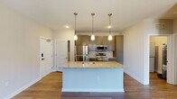 This single-story unit on Ileagnes Road in Raleigh, NC offers a thoughtfully designed open-concept layout. The kitchen serves as the focal point, showcasing gray shaker-style cabinetry extending to the ceiling, granite countertops, and a full suite of stainless steel appliances including a refrigerator, range, and over-the-range microwave. A decorative patterned tile backsplash adds visual interest behind the cooking area. A large central island with a granite surface and an integrated sink is illuminated by three gold-toned pendant lights, creating a natural gathering space that connects seamlessly to the living area. Wood-look flooring runs throughout the main living spaces, lending warmth and continuity. The bedroom features plush carpet, a ceiling fan with light fixture, and a window that allows natural light to filter in. A dedicated laundry area houses a stacked washer and dryer unit, adding everyday convenience. The bathroom includes a full tub-shower combination with chrome fixtures and a curved shower rod. Neutral wall tones and clean white trim throughout create a bright, cohesive aesthetic well-suited to a variety of furnishing styles.