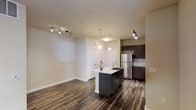 This single-floor space showcases a thoughtfully designed open-concept layout that seamlessly integrates the kitchen and living areas. The rich, dark hardwood flooring runs continuously throughout, providing a cohesive and grounded aesthetic against the neutral, light-toned walls.

The kitchen serves as a central focal point, anchored by a freestanding island with a light stone countertop and an undermount sink. Dark espresso cabinetry lines the perimeter walls, offering generous storage, while a stainless steel refrigerator and a classic subway tile backsplash contribute to the contemporary feel. Two elegant pendant lights hang above the island, complementing the additional track lighting installed in the adjoining living area, which provides flexible, directed illumination throughout the space.

The open living area offers a generous sense of space, benefiting from the flowing layout and consistent flooring. Two paneled doors are visible at the rear of the unit, suggesting access to additional rooms or closet storage. Overall, the space presents a clean, modern aesthetic with a functional and well-considered design.