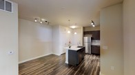 This single-floor space showcases a thoughtfully designed open-concept layout that seamlessly integrates the kitchen and living areas. The rich, dark hardwood flooring runs continuously throughout, providing a cohesive and grounded aesthetic against the neutral, light-toned walls.

The kitchen serves as a central focal point, anchored by a freestanding island with a light stone countertop and an undermount sink. Dark espresso cabinetry lines the perimeter walls, offering generous storage, while a stainless steel refrigerator and a classic subway tile backsplash contribute to the contemporary feel. Two elegant pendant lights hang above the island, complementing the additional track lighting installed in the adjoining living area, which provides flexible, directed illumination throughout the space.

The open living area offers a generous sense of space, benefiting from the flowing layout and consistent flooring. Two paneled doors are visible at the rear of the unit, suggesting access to additional rooms or closet storage. Overall, the space presents a clean, modern aesthetic with a functional and well-considered design.