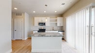 Located at 810–816 Robbie View in Colorado Springs, CO, this single-floor unit offers a clean, contemporary design with an open and functional layout. The kitchen serves as the centerpiece of the living space, showcasing a large center island topped with striking green-veined granite countertops, white shaker-style cabinetry, open shelving, and a full suite of stainless steel appliances including a gas range, microwave, and dishwasher. Pendant lighting and recessed ceiling lights illuminate the space, while light hardwood-style flooring extends throughout the main areas, adding warmth and continuity. A sliding glass door with vertical blinds brings in natural light and provides access to an outdoor area. The hallway connects the main living space to the bathroom and additional rooms, maintaining a cohesive neutral color palette throughout. The bathroom features a white vanity with granite countertop, a framed mirror, and a full tub-shower combination with tiled surround. A generously sized walk-in closet with wire shelving systems and carpeted flooring provides ample storage. A ceiling fan is present in one of the rooms, adding comfort and practicality. Overall, the unit presents a bright, move-in ready atmosphere with modern finishes throughout.