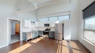 Located at 4545 Forest Park Ave in St. Louis, Missouri, this single-floor loft-style unit showcases an industrial-modern design characterized by soaring ceilings with exposed ductwork and generous natural light throughout. Rich hardwood-style flooring runs through the main living areas, anchoring the open, airy layout.

The living area is expansive, benefiting from oversized windows and a sliding glass door that opens to a private balcony with city views. A ceiling fan adds comfort, while track lighting along the hallway provides functional illumination throughout.

The kitchen is well-appointed with two-tone cabinetry—white upper cabinets paired with gray lower cabinets—set against a classic white subway tile backsplash. A full suite of stainless steel appliances includes a refrigerator, range, microwave, and dishwasher, complemented by ample counter space and a stainless steel sink.

A light-filled hallway connects the private areas of the unit, including a carpeted bedroom featuring a sliding closet and clerestory windows. The bathroom offers a gray vanity with a white countertop, and a separate walk-in shower enclosed by a glass door and finished with white subway tile.