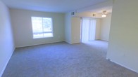 This single-floor space features an open and airy layout with wall-to-wall carpeting throughout the main living area, providing a comfortable and cohesive feel. The space is bathed in natural light, courtesy of a large sliding window on one wall that offers views of the surrounding greenery. A set of vertical blinds frames a sliding glass door, which connects the interior to an outdoor area and further enhances the brightness of the room. The neutral, light-toned walls complement the soft carpeting, creating a clean and understated aesthetic. A ceiling-mounted light fixture is visible in an adjoining area, suggesting the space extends beyond the main living zone. Overhead HVAC vents are integrated into the ceiling, ensuring climate comfort throughout. The open floor plan allows for flexible furniture arrangements, making the space highly adaptable for various living configurations. Overall, the space presents a functional and straightforward design with ample natural light, a neutral color palette, and practical features suited for everyday living.