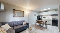 This single-floor unit offers a thoughtfully designed layout that maximizes functionality and comfort. The living area is bright and inviting, featuring light wood-style flooring, a textured ceiling, and a wall-mounted air conditioning unit. Large windows fitted with horizontal blinds allow natural light to fill the space while providing views of the surrounding greenery outdoors. A sectional sofa, glass-top coffee table, and wall art contribute to a clean, contemporary aesthetic.

The kitchen is a standout feature, showcasing gray cabinetry, a classic white subway tile backsplash, and a skylight that bathes the space in natural light. Black appliances, including a refrigerator and gas range, complement the modern design, while ample upper and lower cabinet storage ensures practicality. A dining area sits adjacent to the kitchen, comfortably accommodating a four-person table.

The bedroom is a calm retreat, offering light flooring, neutral-toned walls, and generous storage with a multi-drawer wooden dresser. The full bathroom is clean and well-appointed, featuring a tub-shower combination with white tile surround, a single vanity with a large mirror, and matching light flooring that ties the space together cohesively throughout.
