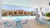 Located at 3900 Dacoma St in Houston, Texas, this outdoor amenity area offers a resort-inspired environment designed for relaxation and social gathering. At its centerpiece is a generously sized swimming pool with clear blue water, framed by smooth stone tile decking and lined with rows of contemporary white lounge chairs. Tall palms and lush tropical plantings add a verdant backdrop, enhancing the open-air atmosphere under abundant Texas sunshine.

A covered pavilion anchors one end of the pool area, constructed with light brick masonry and supported by sturdy columns. The pavilion features ceiling fans for ventilation, a wall-mounted flat-screen television, and built-in outdoor kitchen or grill stations — making it well-suited for communal dining and entertaining. Modern outdoor dining tables and chairs furnish the space throughout.

Adjacent covered patios provide additional shade and seating, outfitted with vibrant teal bistro sets that add a lively pop of color. Wrought iron fencing defines the perimeter of the pool deck, while carefully maintained garden beds with ornamental grasses and shrubs add textural landscaping detail throughout. The multi-story residential building rises on multiple sides of the courtyard, with private balconies overlooking the pool below, creating an enclosed, community-centered outdoor retreat.