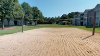 This community amenity space offers a well-rounded recreational and social environment designed to bring residents together. Outdoors, a full-size sand volleyball court provides a dedicated space for active play, set against a backdrop of lush green trees and open sky. Surrounding greenery and nearby structures create a pleasant, park-like atmosphere for outdoor gatherings and friendly competition.

The interior clubhouse is equally impressive, characterized by soaring vaulted ceilings adorned with striking exposed wooden beams that lend a warm, rustic-modern charm. Large multi-pane windows flood the space with abundant natural light, enhancing the bright, airy ambiance throughout. The main lounge area features a cozy fireplace, stylish tufted seating in rich jewel tones, and natural wood accent tables, creating an inviting setting for relaxation and socializing.

Adjacent to the lounge, a compact kitchen with dark cabinetry and stainless steel appliances supports events and gatherings with practical convenience. A dedicated business center offers workstation desks and ergonomic seating beneath the same dramatic vaulted ceiling, making it a functional yet refined space for residents to work or study. Hardwood-style flooring and wainscoting paneling tie the interior together, delivering a cohesive, polished aesthetic throughout.